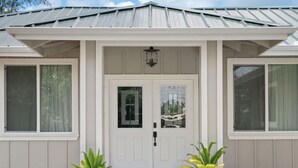 A two-story house with a white front door, large windows, and a small porch area with potted plants. - Gorgeous Gated Retreat Near Ocean w/ Pool, Deck, Firepit on 1+ acre! (Keaau)