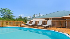 A swimming pool with lounge chairs and a wooden deck, surrounded by a wooden fence and trees. - Gorgeous Gated Retreat Near Ocean w/ Pool, Deck, Firepit on 1+ acre! (Keaau)