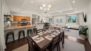 A spacious living area with a dining table set for six, a kitchen with wooden cabinets, and a large window providing natural light. - Gorgeous Gated Retreat Near Ocean w/ Pool, Deck, Firepit on 1+ acre! (Keaau)