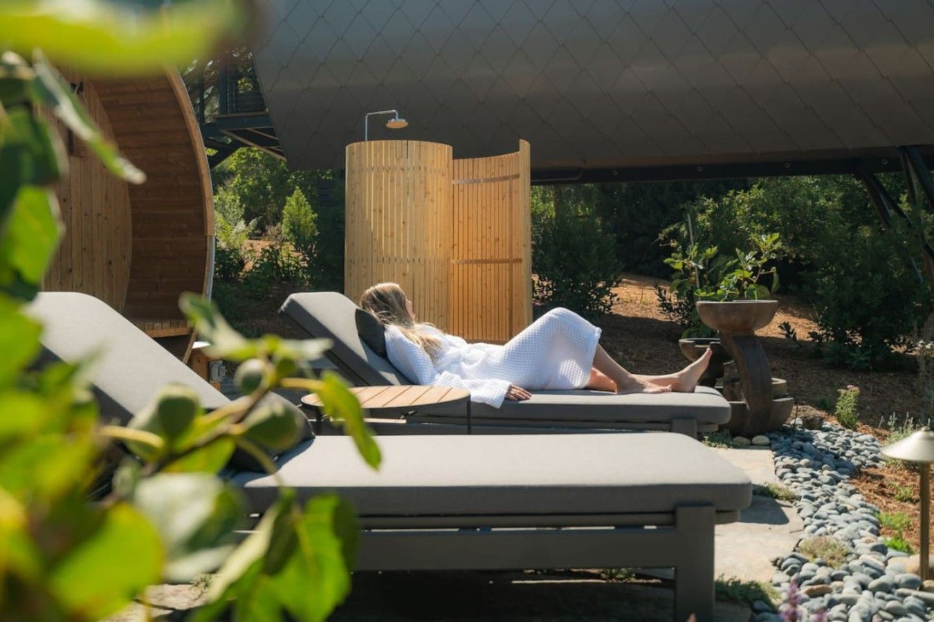 Eine Person entspannt sich auf einer Liege unter einer Holzpergola, umgeben von viel Grün und einem Kiesweg.