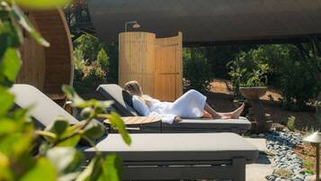 Eine Person entspannt sich auf einer Liege unter einer Holzpergola, umgeben von viel Grün und einem Kiesweg.