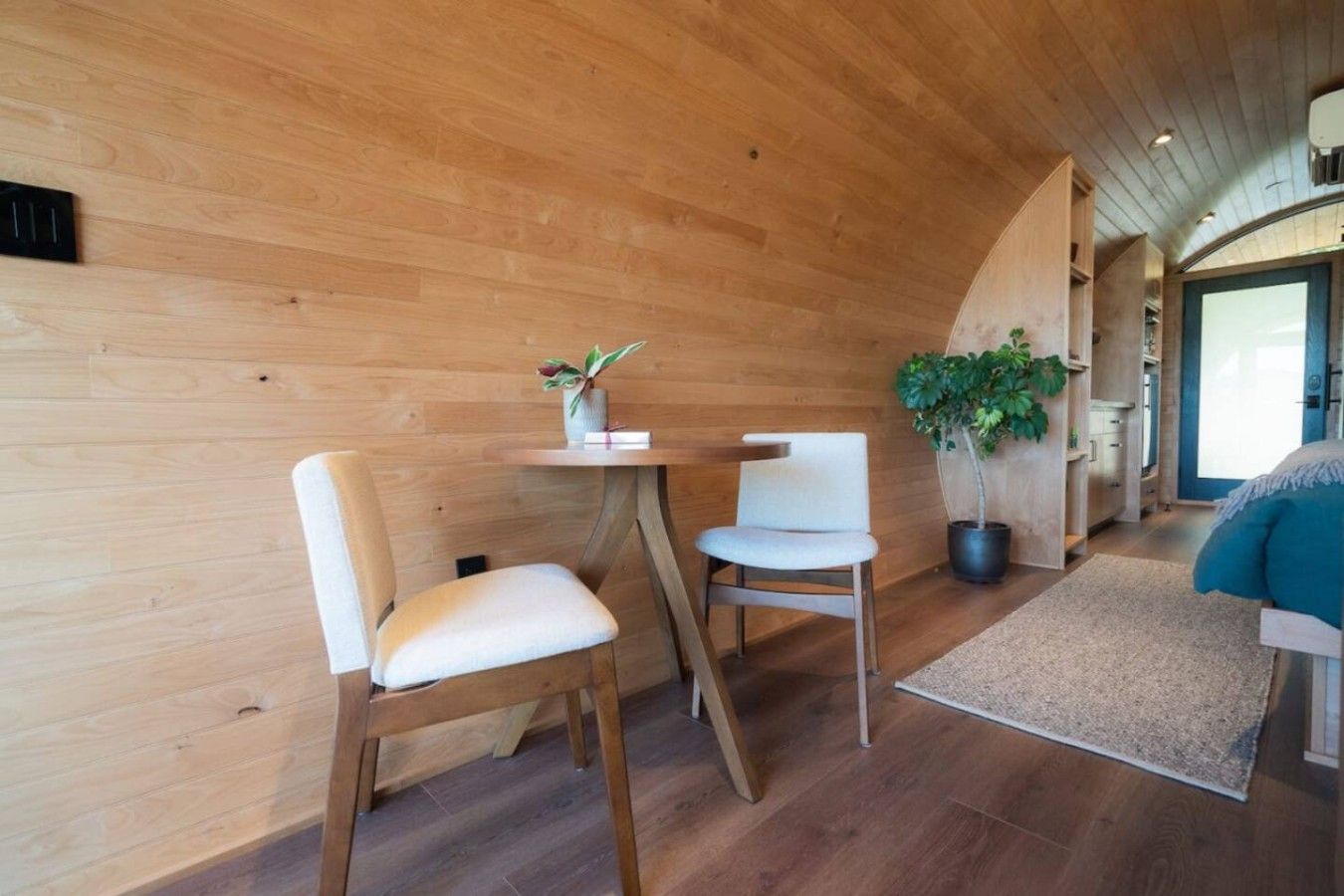 Ein Zimmer mit Holzwänden, einem runden Tisch, zwei Stühlen, einer Topfpflanze und einem Bett mit blauer Decke.