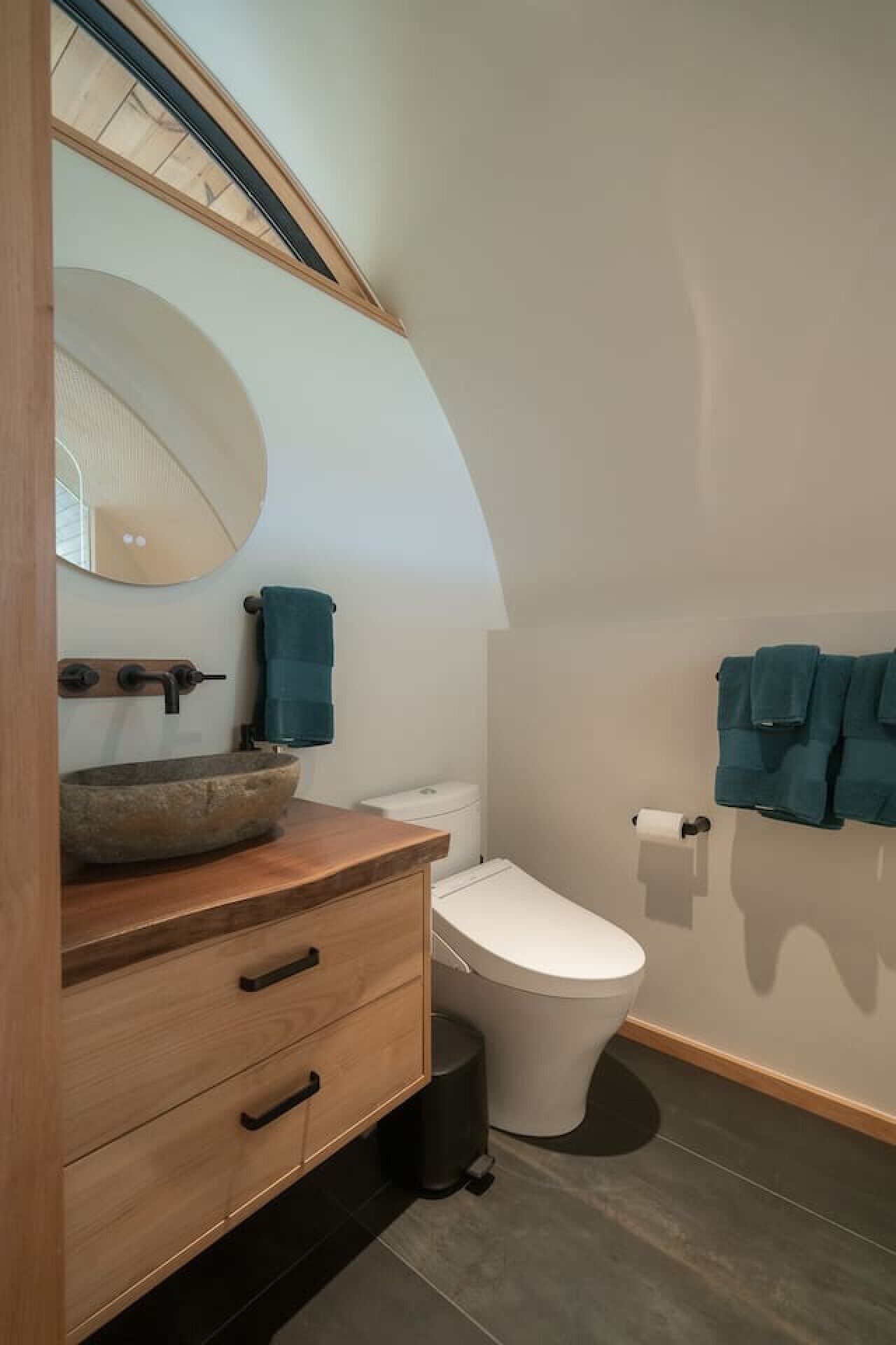 A bathroom with a wooden vanity, a round mirror, a toilet, and a towel rack.