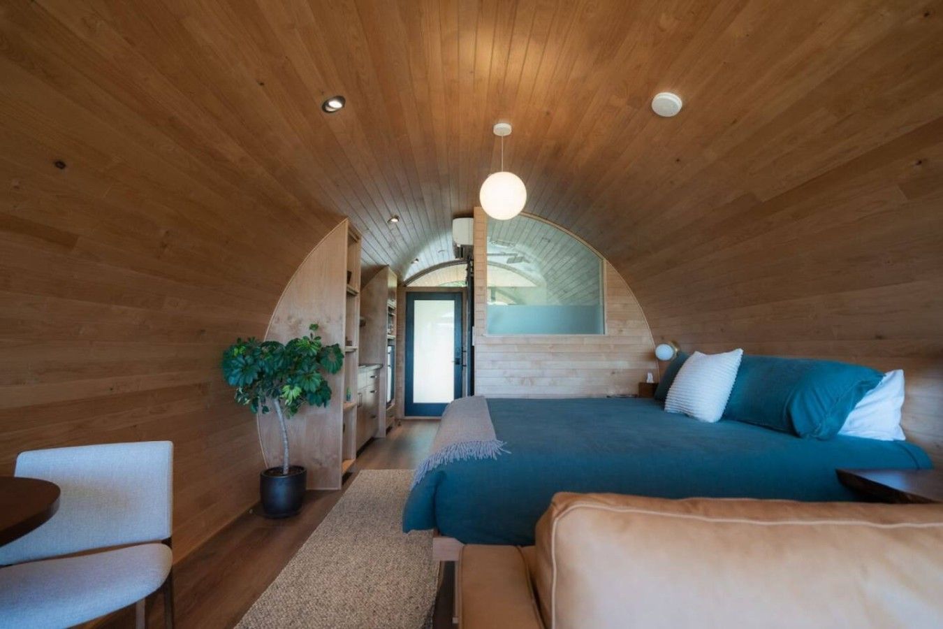 Ein gemütliches Schlafzimmer im Dachgeschoss mit gewölbter Decke, einem Bett mit blauer Bettwäsche, einem Topf mit einer Pflanze und einer kleinen Sitzecke.