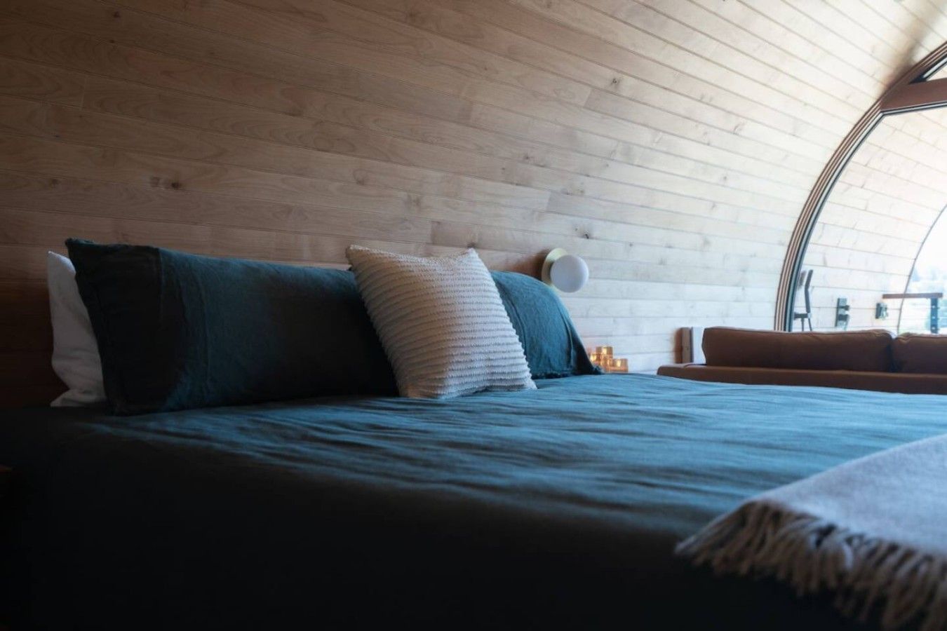 Un dormitorio con techo de madera, cama con ropa oscura y una lámpara redonda fijada a la pared.