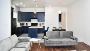 A modern living room with a grey sofa, a white coffee table, and a kitchen area with dark cabinets and a blue island.
