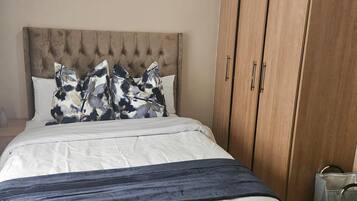 A bedroom with a tufted headboard, a bed with white bedding and floral pillows, a dark blue throw, and a wooden wardrobe with a laundry bag on the floor.