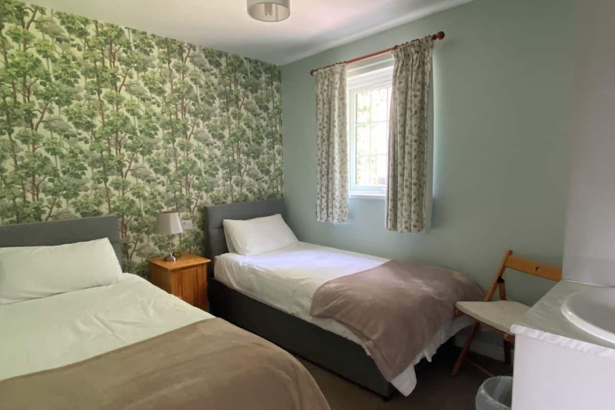 Une chambre avec deux lits, une fenêtre avec des rideaux, un ventilateur de plafond et un mur décoré d’un motif d’arbre.
