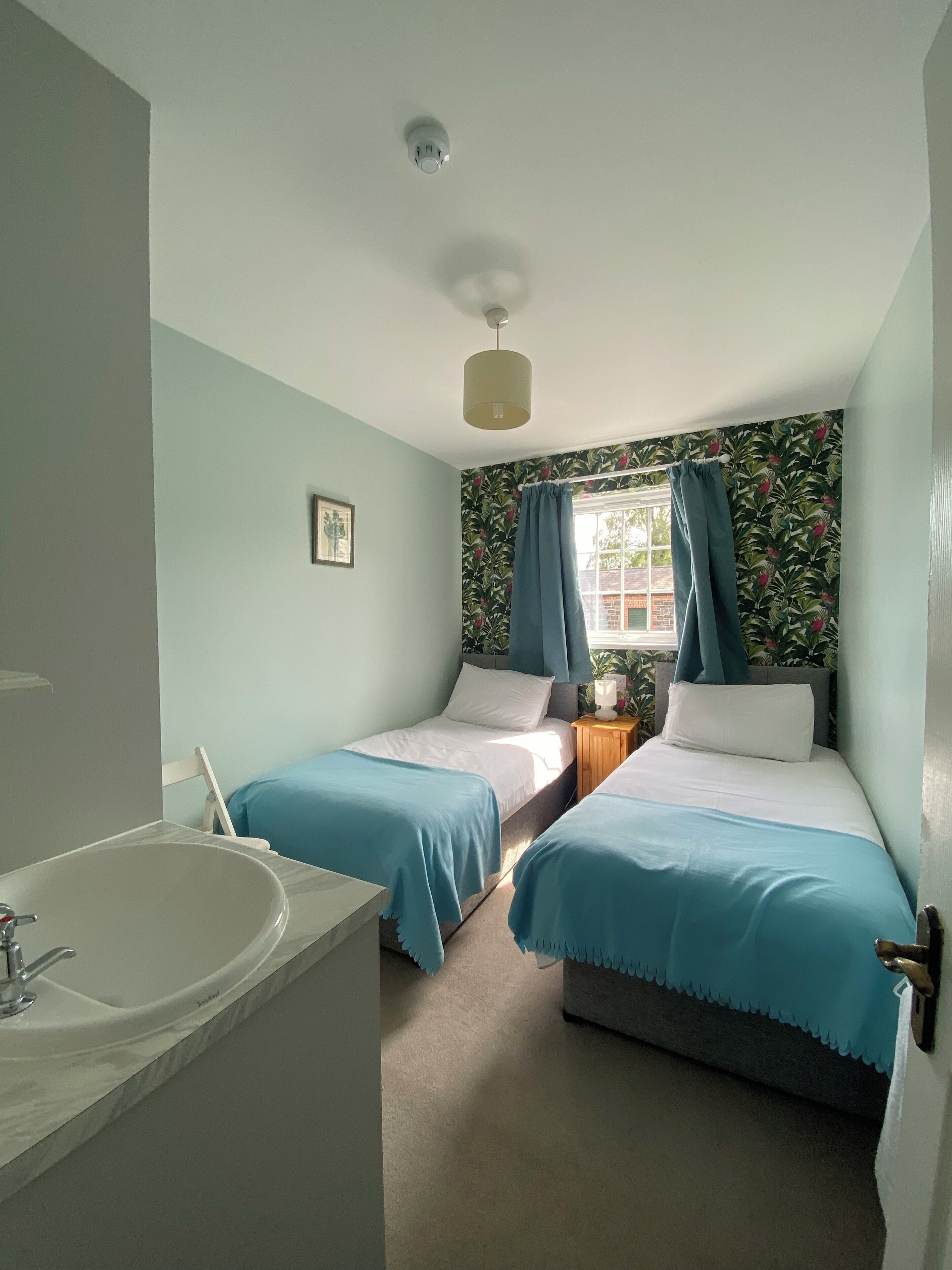 Une chambre avec deux lits simples, une salle de bain avec un lavabo et une fenêtre avec des rideaux.