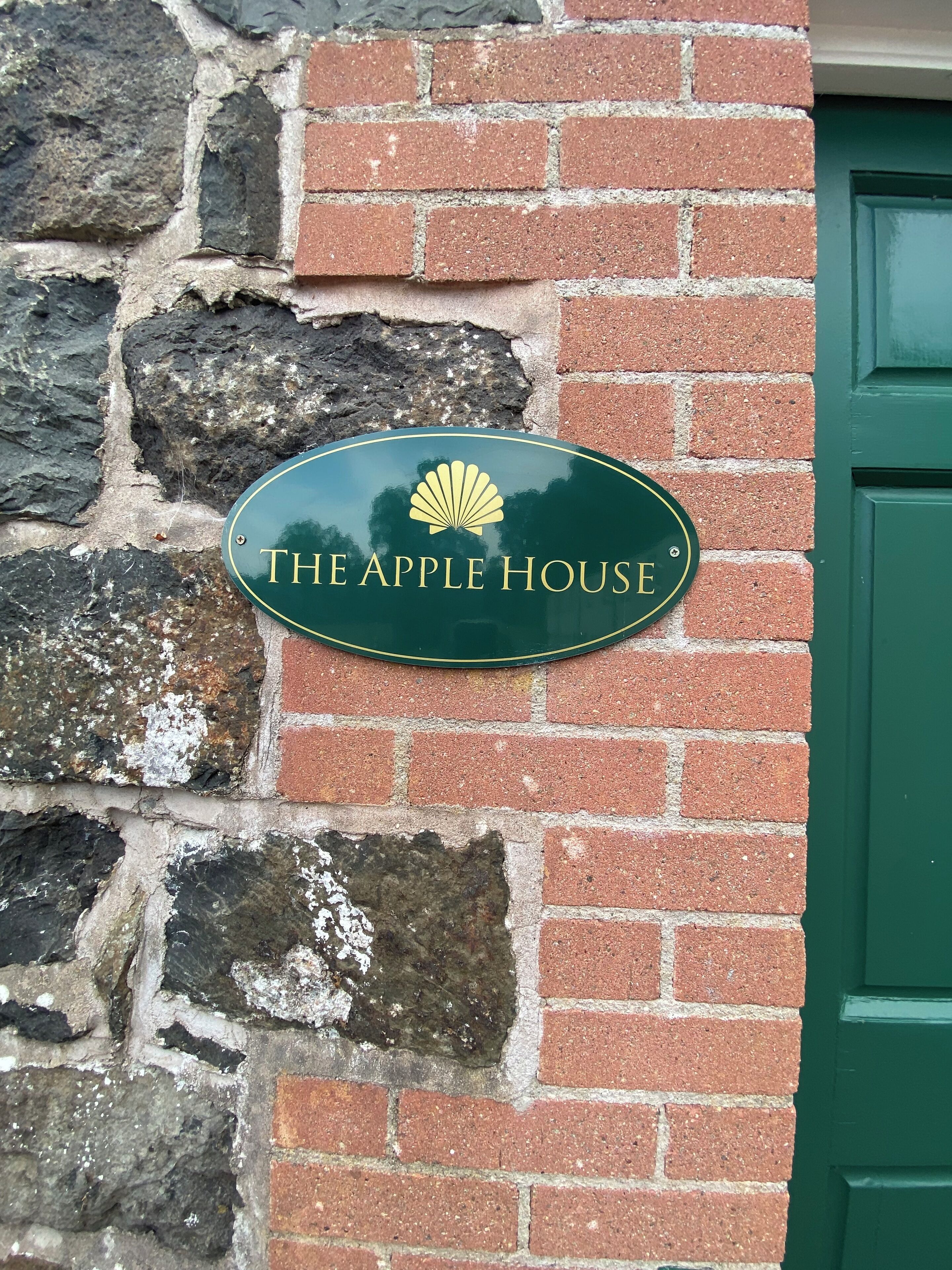 Un letrero verde con un logotipo de caracola y el texto "THE APPLE HOUSE" sobre una pared de ladrillo.