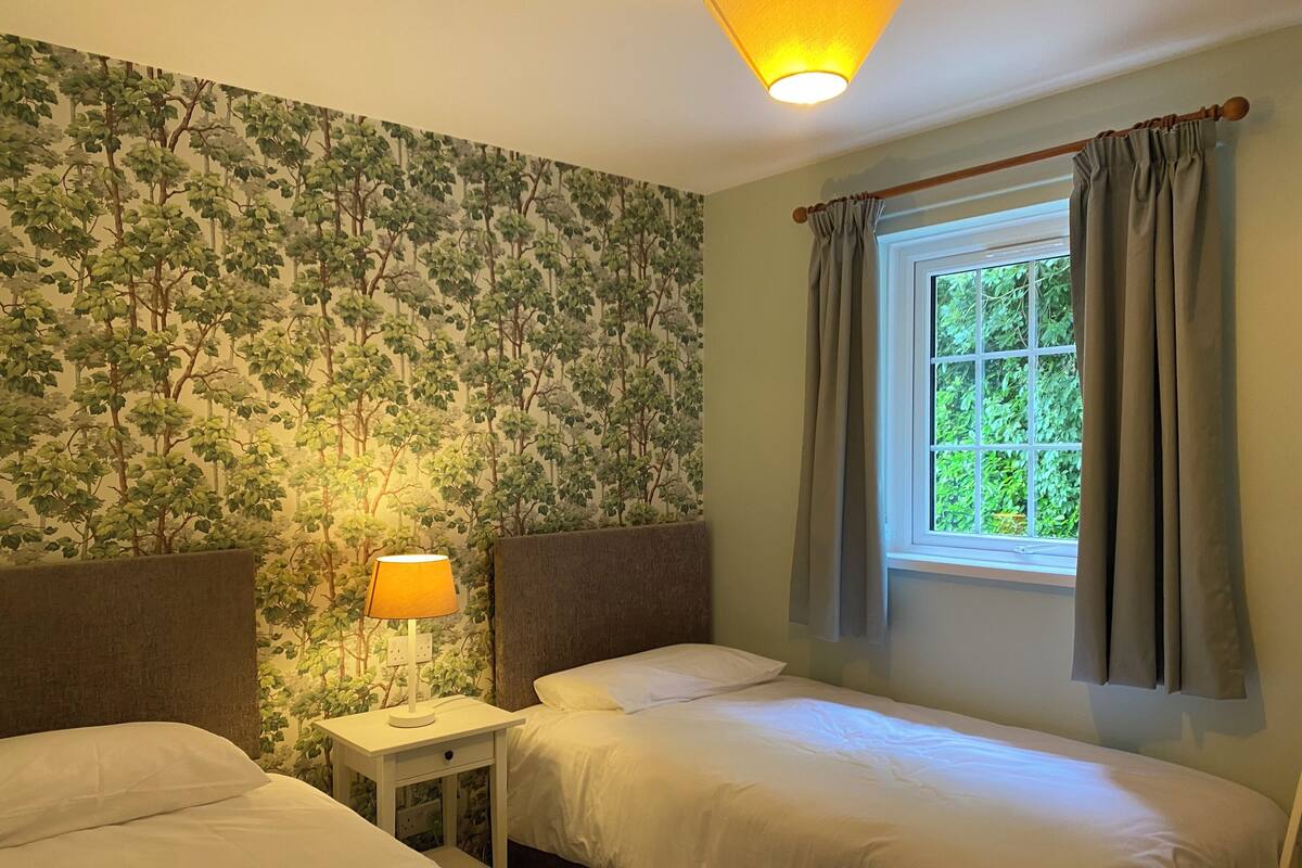 Une chambre avec deux lits, une table de chevet, une lampe, une fenêtre avec des rideaux et un mur recouvert d’un papier peint à motifs de feuilles.