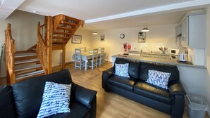 A modern living room with a black leather sofa, a wooden staircase, a dining area with a white table and chairs, and a kitchen with white cabinets and appliances. - Ballyscullion Park Courtyard Cottages (Magherafelt)