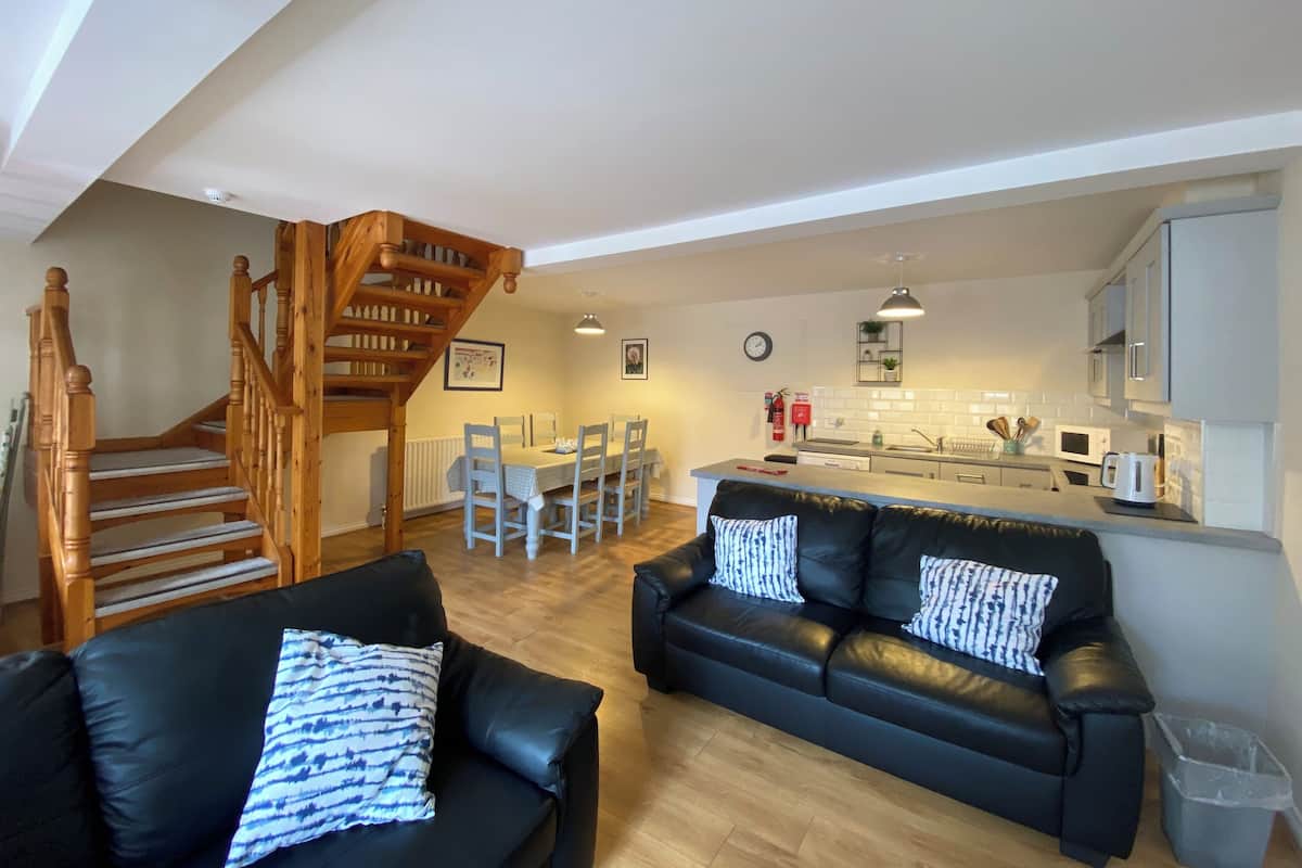 Un salon moderne avec un canapé en cuir noir, un escalier en bois, un coin repas avec une table et des chaises blanches, et une cuisine avec des meubles et des appareils électroménagers blancs.