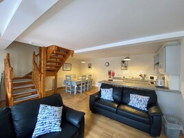 Uma sala de estar moderna com sofá de couro preto, escada de madeira, área de jantar com mesa e cadeiras brancas e cozinha com armários e eletrodomésticos brancos.