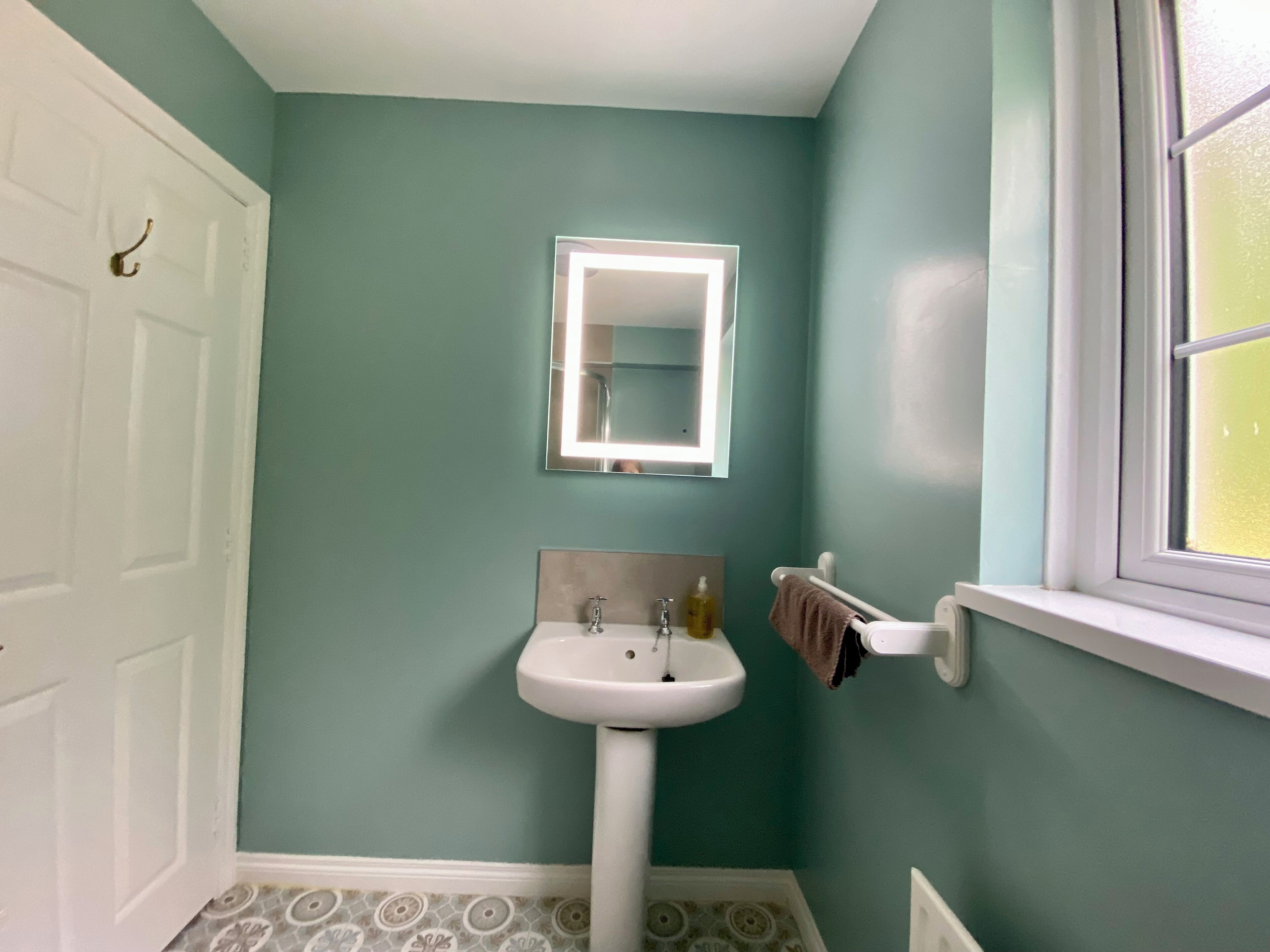 Une salle de bain aux murs de couleur turquoise, avec un lavabo sur pied blanc, un miroir avec un éclairage intégré et une fenêtre à vitres dépolies.