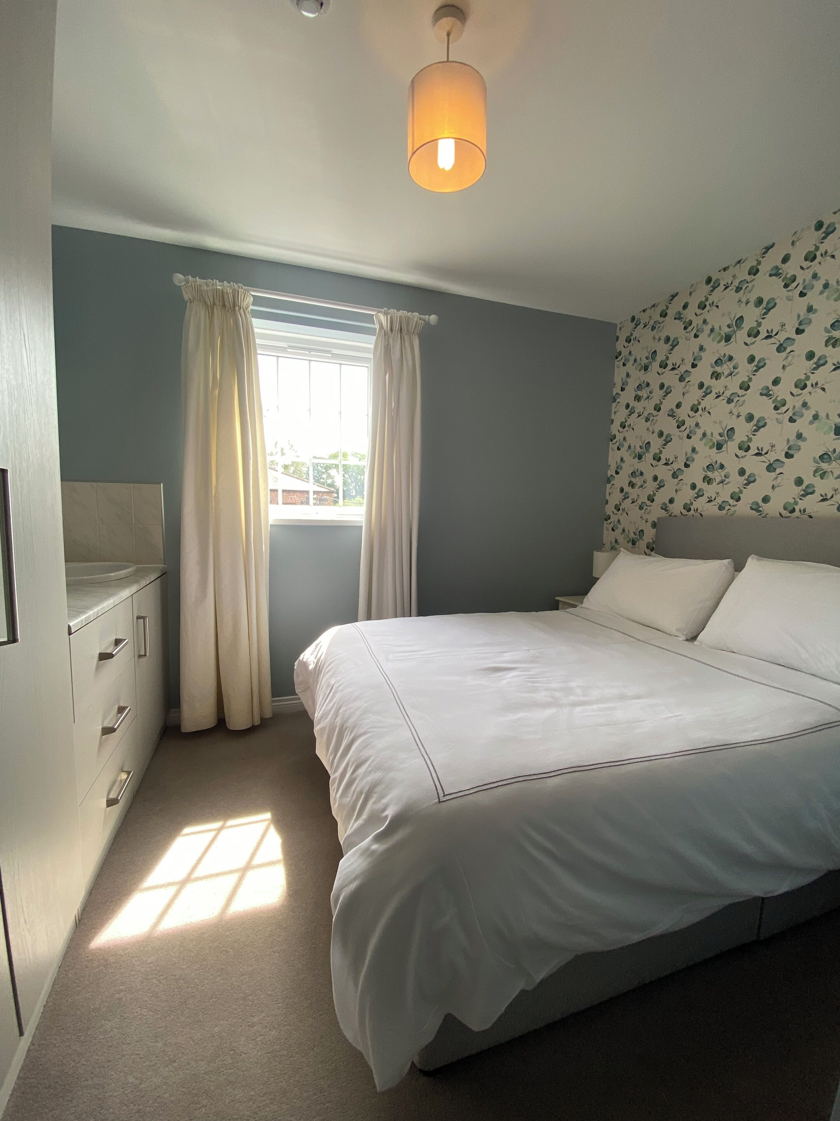 Een slaapkamer met een bed, een raam met gordijnen, een nachtkastje en een bloemenprint op de muur.