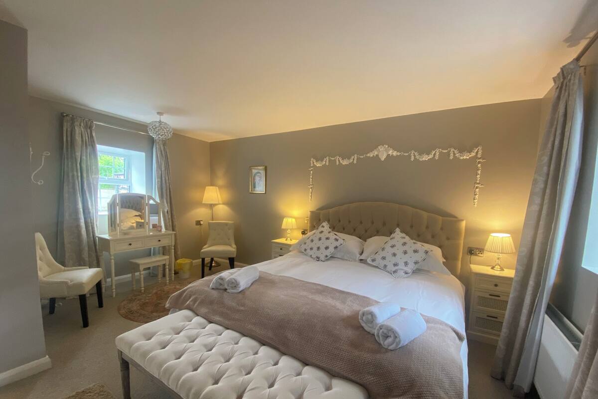 Une chambre avec un grand lit, deux tables de chevet avec des lampes, un bureau avec une chaise et un banc.