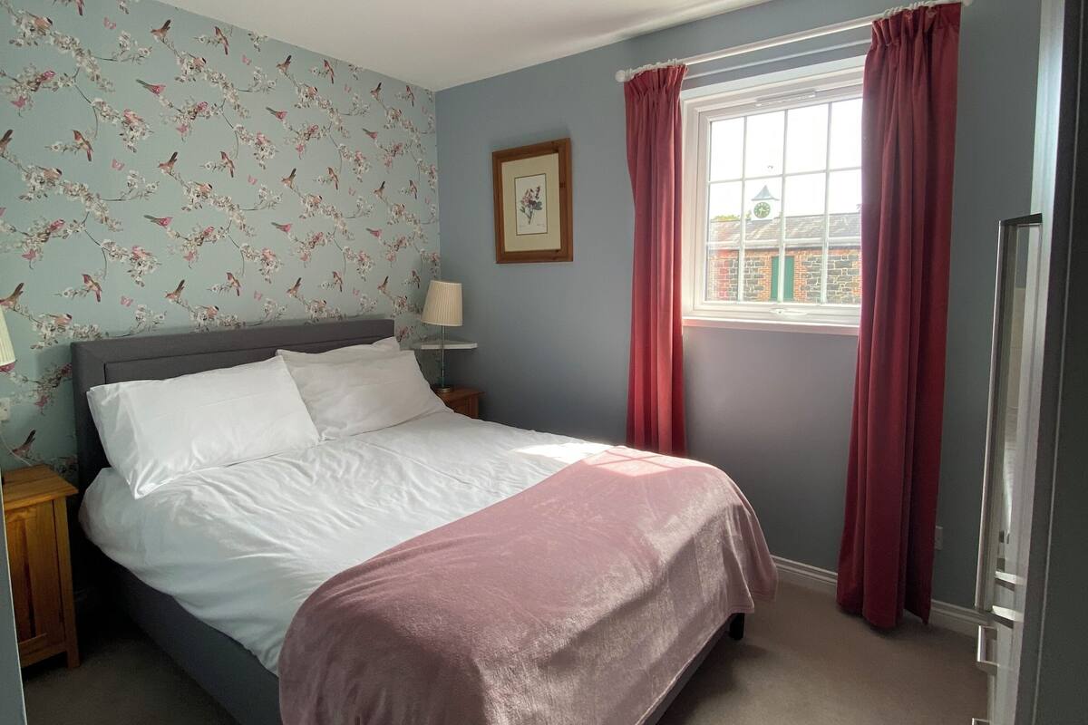 Une chambre à coucher comprenant un lit, une table de chevet, une fenêtre avec des rideaux, une horloge murale et un ventilateur de plafond.