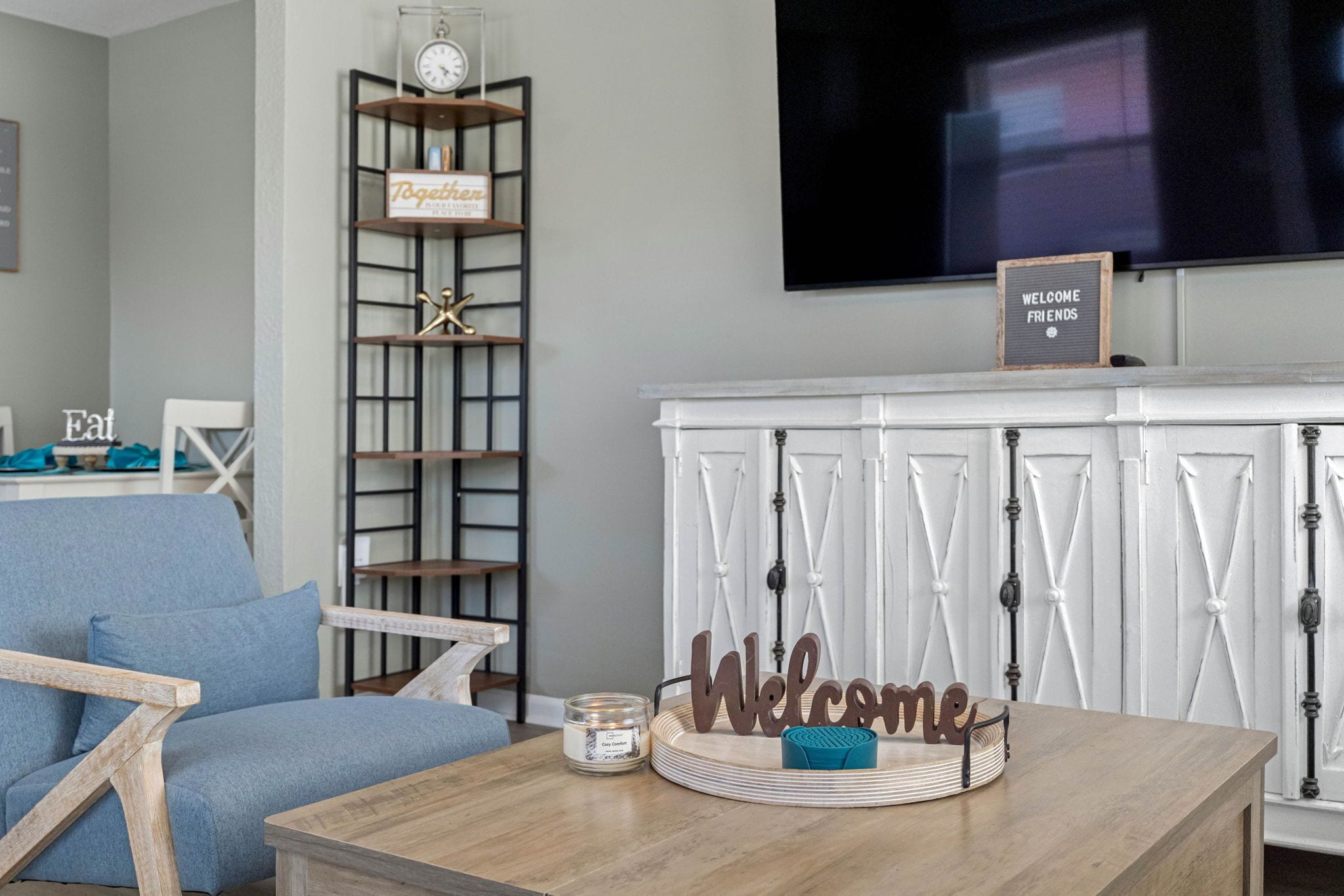 Un soggiorno con un divano blu, un tavolino in legno con una candela e un cartello con la scritta 'Welcome'. C'è una TV montata a parete e una libreria con un cartello 'Welcome'.