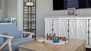 A living room with a blue sofa, a wooden coffee table with a candle, and a 'Welcome' sign. There is a TV mounted on the wall and a bookshelf with a 'Welcome' sign. - Charming Beach Retreat (St. Pete Beach)