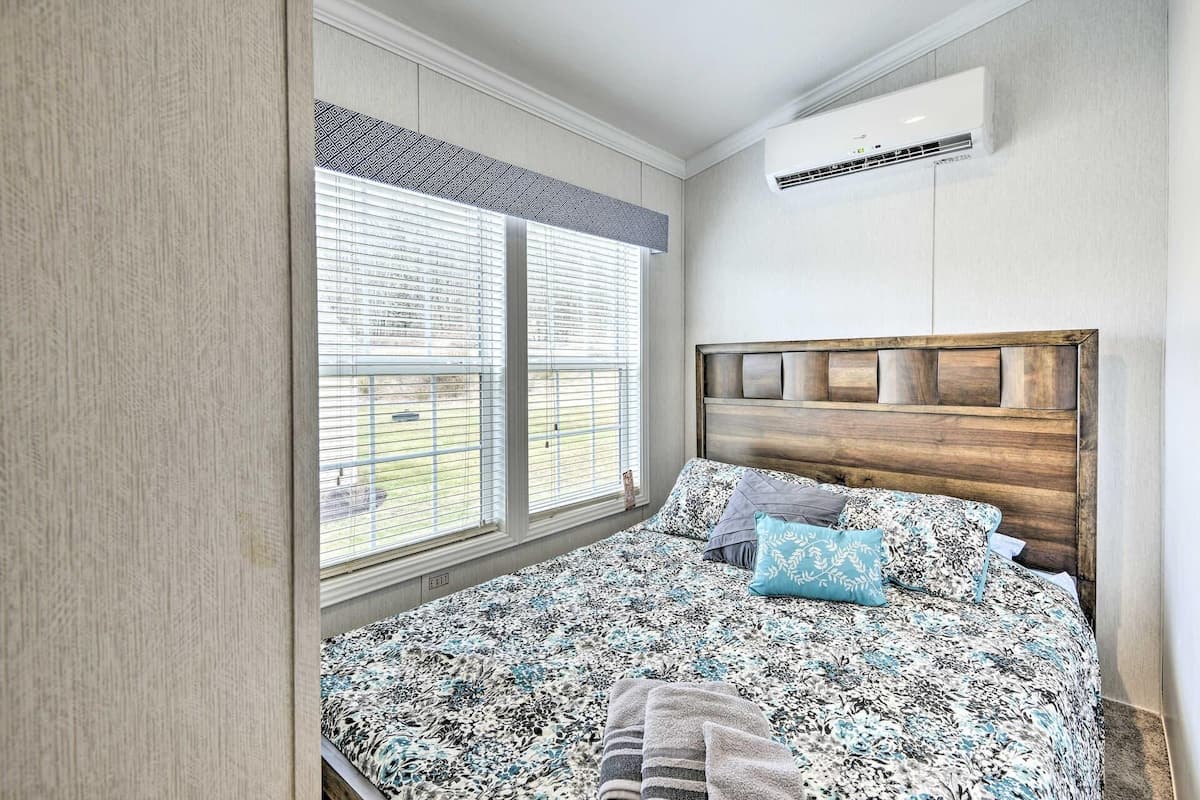 A bedroom with a large window, a bed with patterned bedding, a wooden headboard, and an air conditioning unit on the wall.