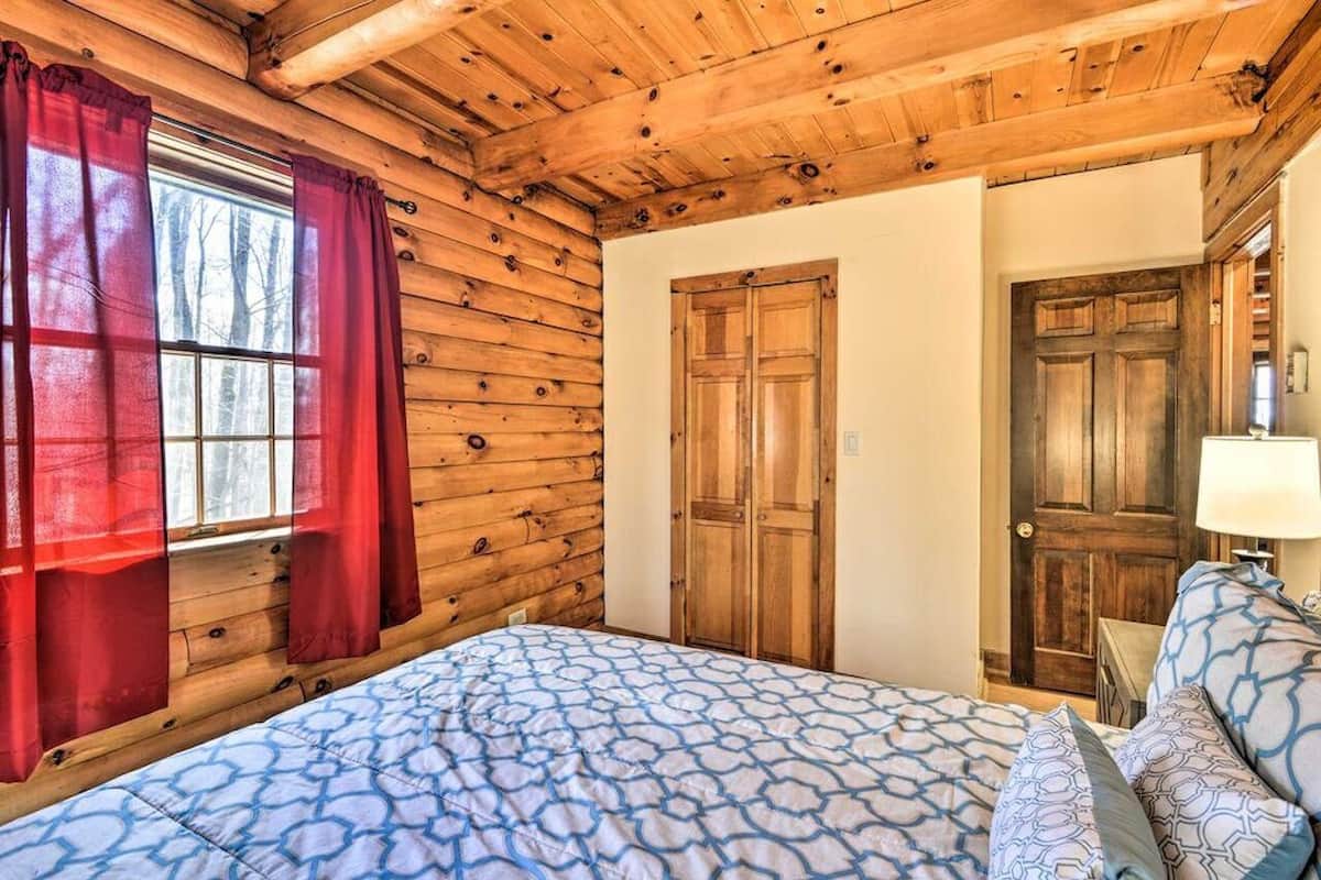 A wooden cabin bedroom with a bed, red curtains, and a wooden door.