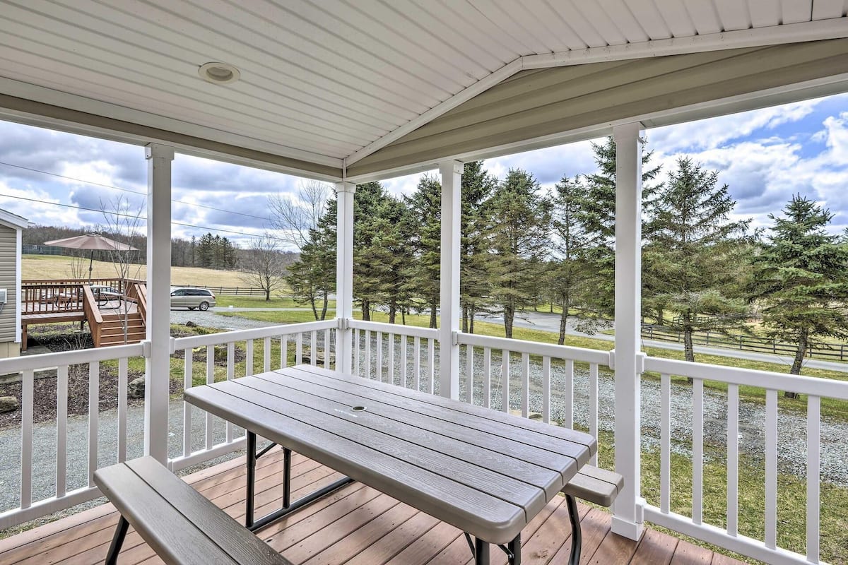 A covered porch with a wooden table and bench, overlooking a driveway and trees.