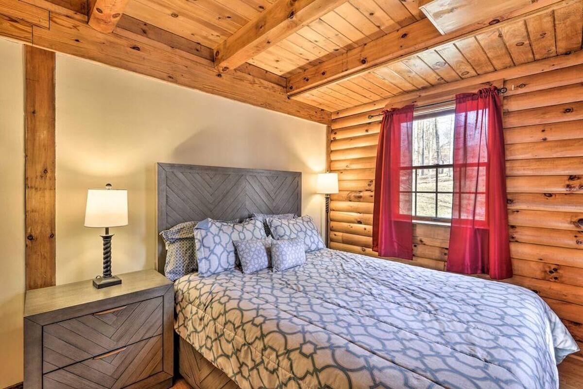 A log cabin bedroom with a bed, two bedside lamps, a window with red curtains, and a wooden ceiling.