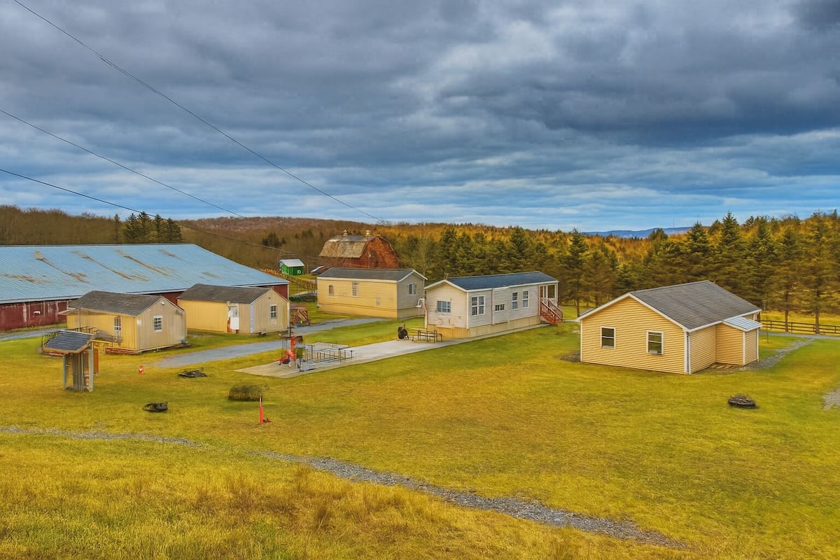 A rural setting with several small buildings, including a barn and residential houses, surrounded by grassy fields and trees.