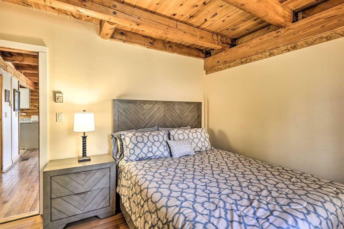 A bedroom with a wooden ceiling, a bed with patterned bedding, a nightstand with a lamp, and a wall clock.