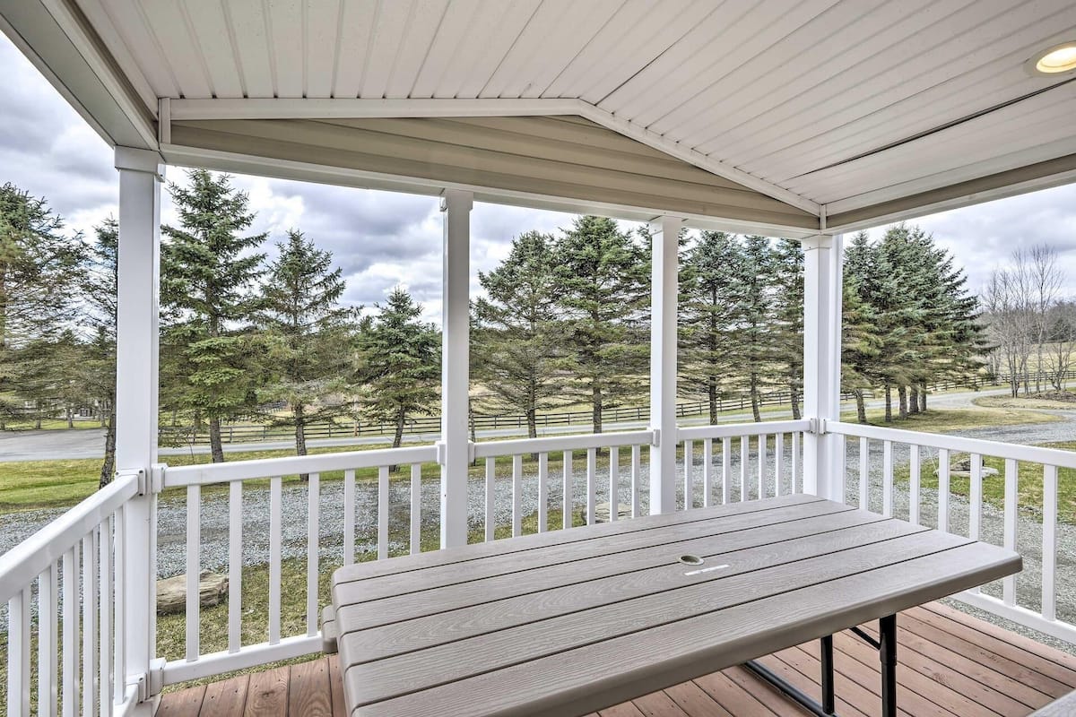 A covered outdoor area with a wooden table and white railings, overlooking a road and trees.