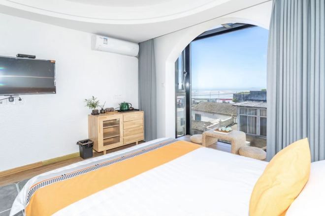 A modern hotel room with a large bed, a TV mounted on the wall, a small desk, and a view of the city through large windows.