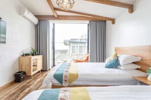 Una habitación de hotel con dos camas, un cabecero de madera, un mueble pequeño de madera, un cesto de basura y vista a la ciudad desde la ventana.