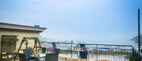 A terrace with a table and chairs, a wooden bench, and a railing overlooking a body of water.