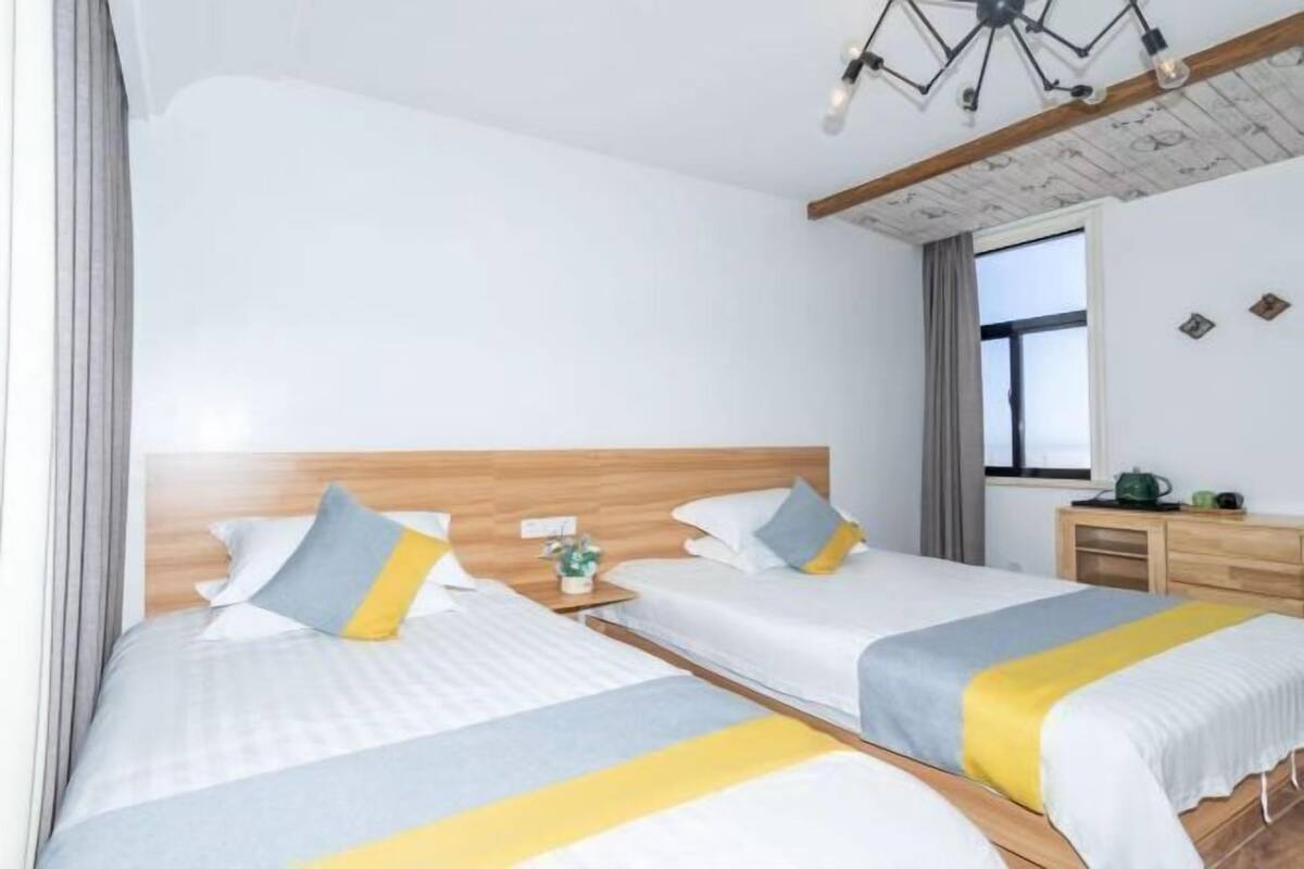 Une chambre d’hôtel avec deux lits, des têtes de lit en bois, une petite table de chevet avec une lampe, une fenêtre avec des rideaux et un luminaire au plafond.