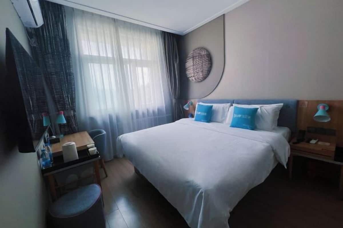 Quarto de hotel com cama, escrivaninha, cadeira, televisão e janela com cortinas.