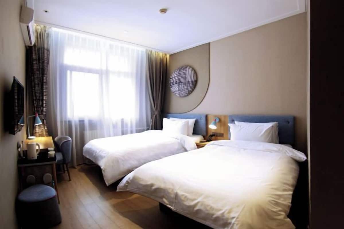 Quarto de hotel com duas camas, uma escrivaninha com luminária, uma cadeira e uma janela grande com cortinas.