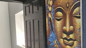 Une porte à la surface texturée et dotée d’une poignée en métal, qui donne sur une pièce dont un mur est bleu et qui contient un grand tableau représentant le visage d’un Bouddha.