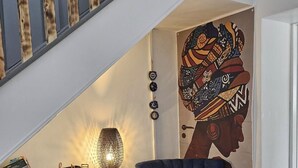 Un salon avec un fauteuil bleu foncé, une bibliothèque remplie de divers ouvrages, une lampe décorative et un mur orné d’une fresque colorée.