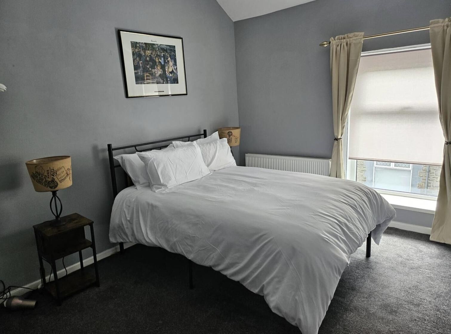 A bedroom with a bed, a framed picture on the wall, a bedside lamp, and a window with curtains.