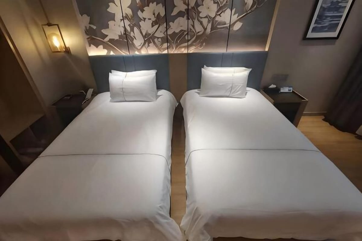 Deux lits simples avec du linge de lit blanc, une lampe fixée au mur et une tête de lit à motifs floraux.