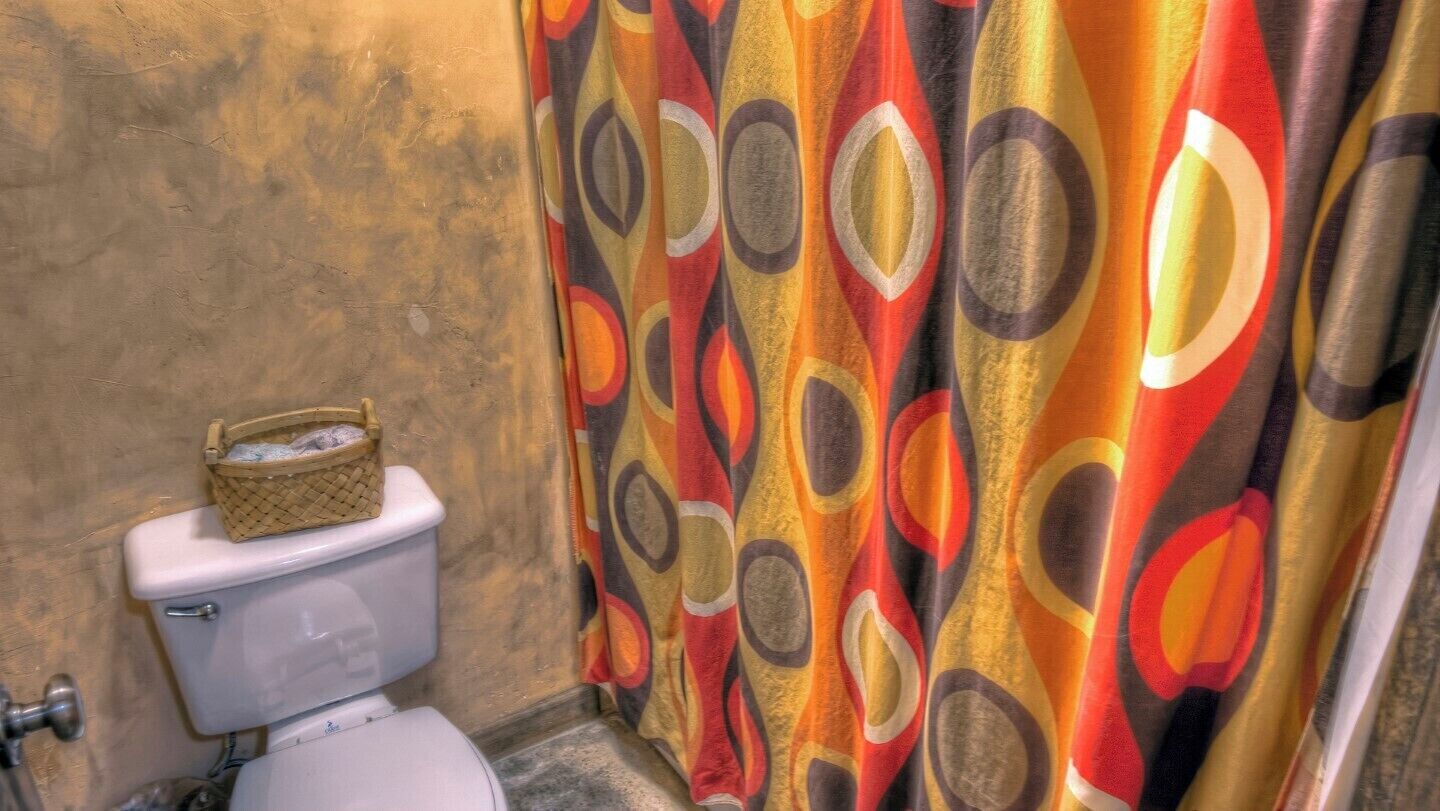 Une salle de bain avec des toilettes, un panier posé sur la cuvette, et un rideau de douche aux couleurs vives.