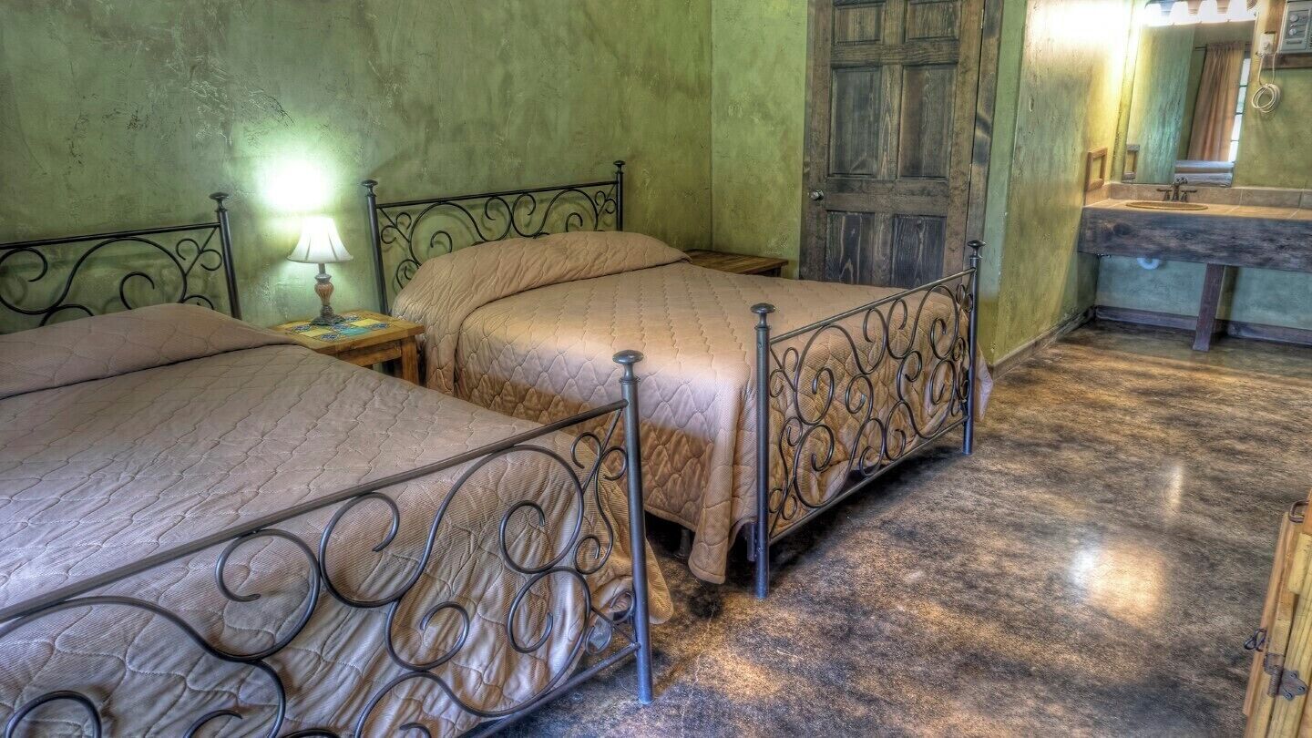Una habitación con dos camas, una puerta de madera, una mesita con l ámpara y un baño con lavabo y espejo.