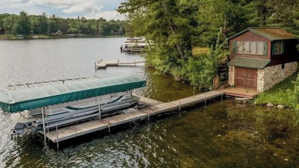 A dock with a covered boat, a small cabin, and a lake with trees in the background.