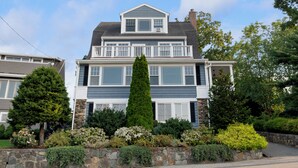 A two-story house with a stone foundation, a large central tree, and a well-maintained garden. - Magnificent Ocean View; Walk to beaches and town (Swampscott)