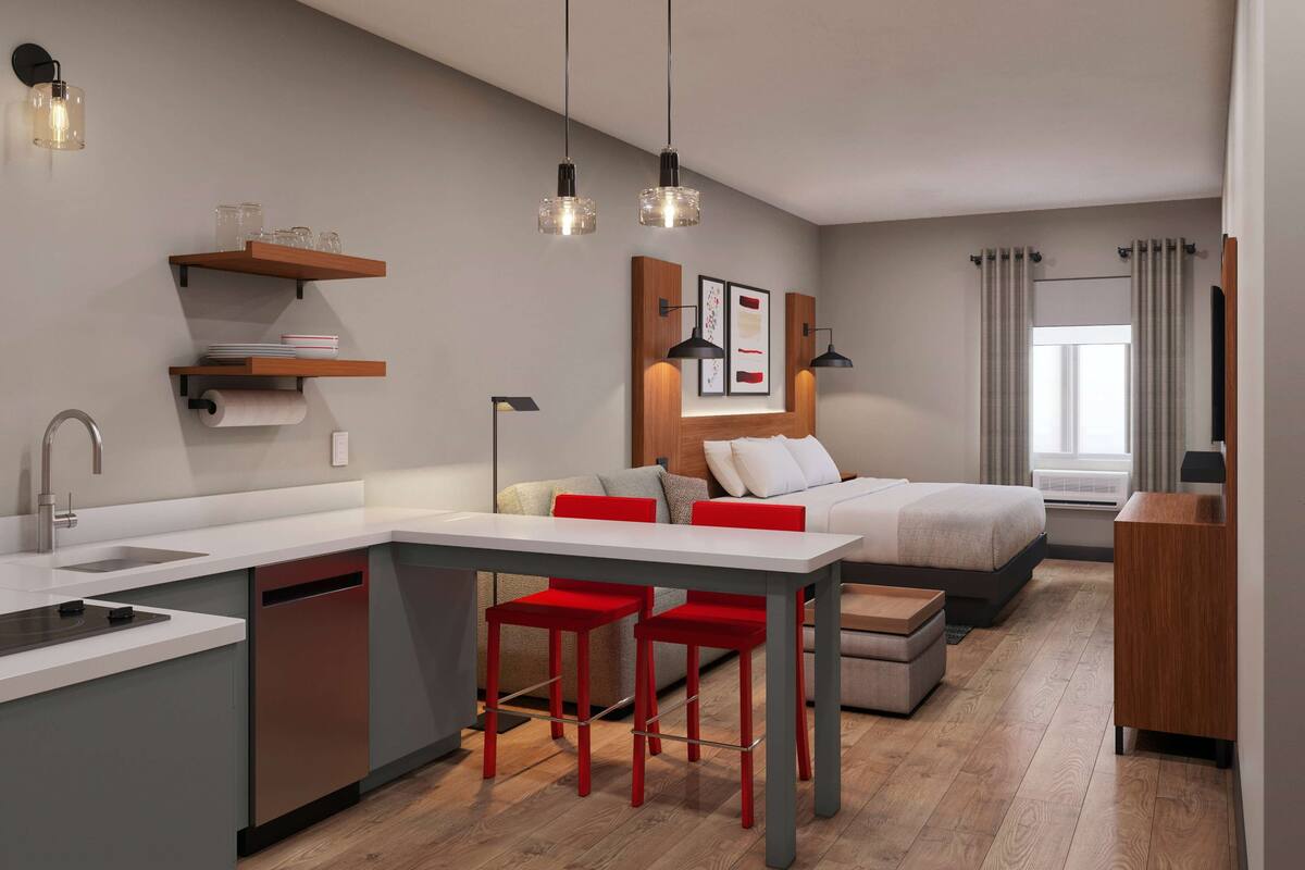 Une cuisine moderne dotée d’un lave-vaisselle intégré, un coin repas avec des chaises rouges, un lit avec une tête de lit et une fenêtre avec des rideaux.