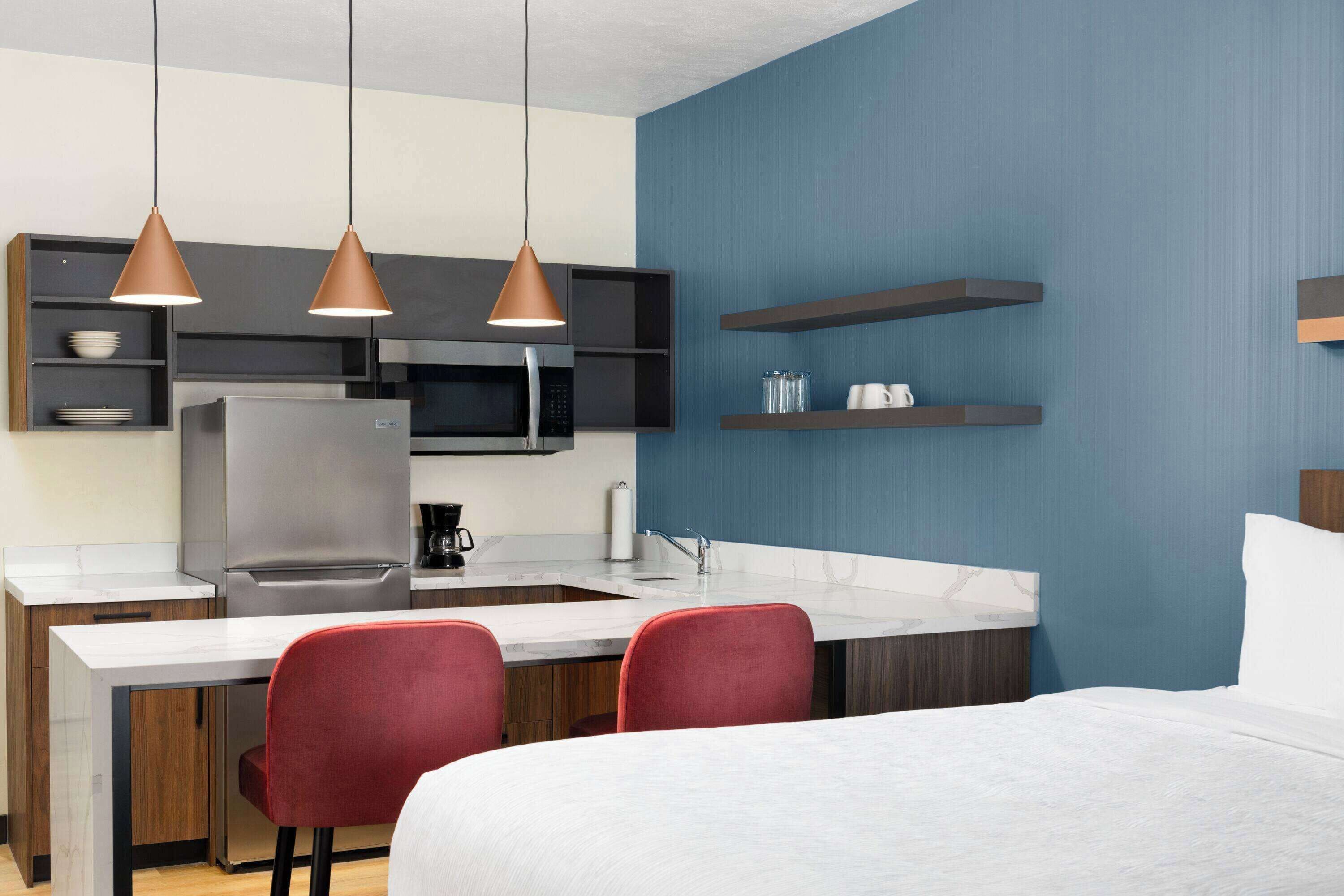 A modern kitchen with a refrigerator, microwave, and coffee maker. There are two red chairs and a bed with white bedding.