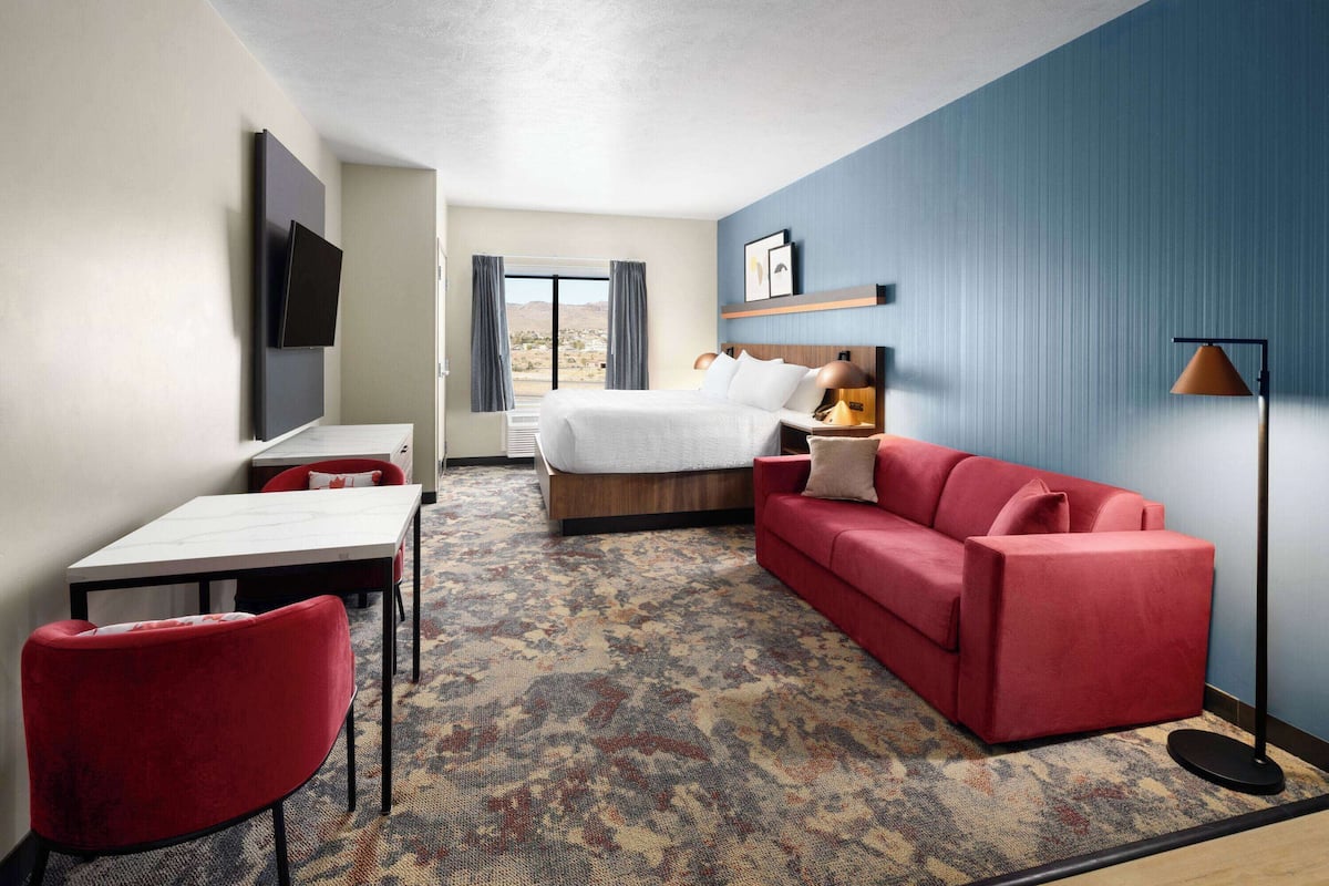 Une chambre d’hôtel avec un lit, un canapé rouge, un bureau, une chaise, un téléviseur fixé au mur et un sol recouvert de moquette.