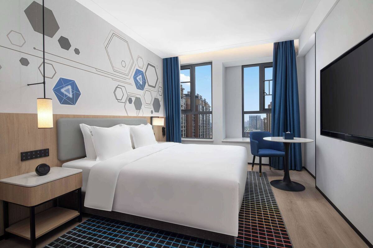 Een moderne hotelkamer met een groot bed, een nachtkastje met een klok, een bureau met een stoel, een televisie en grote ramen met gordijnen.