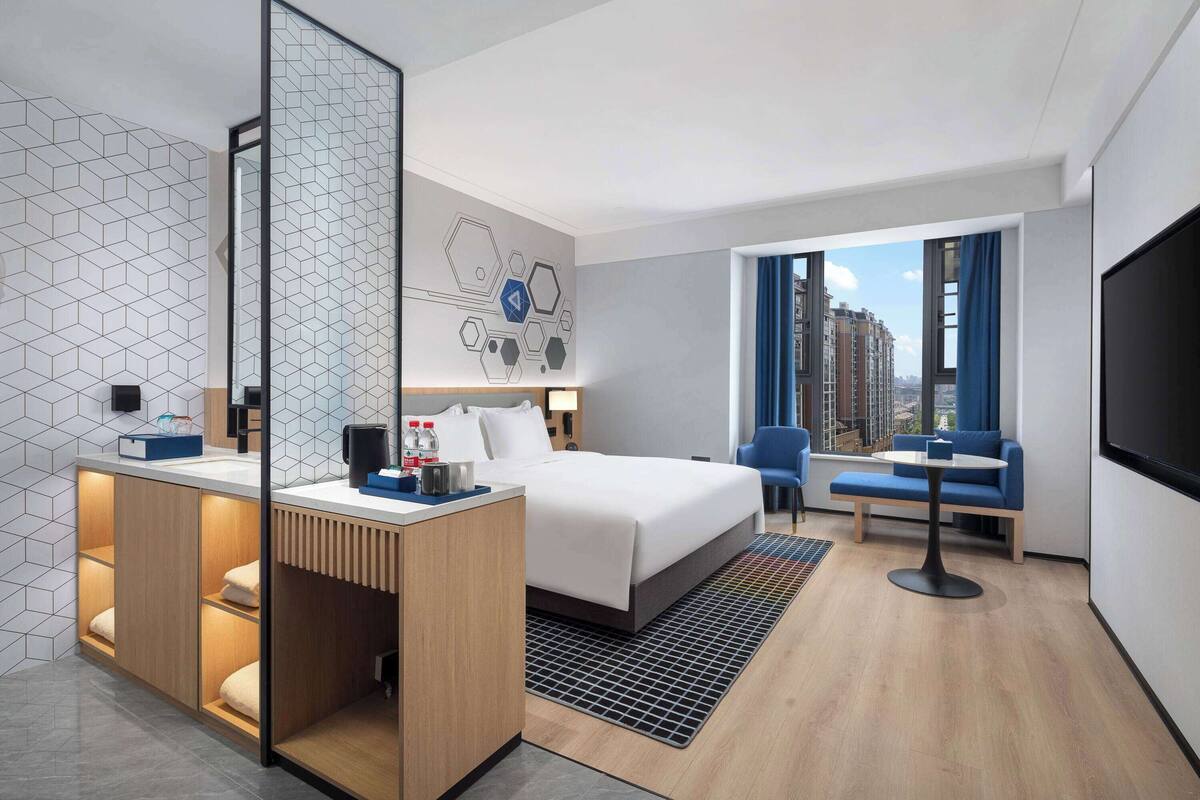 Een moderne hotelkamer met een groot bed, een bureau met spiegel, een zithoek met een bank en stoelen, en een groot raam met uitzicht op de stad.